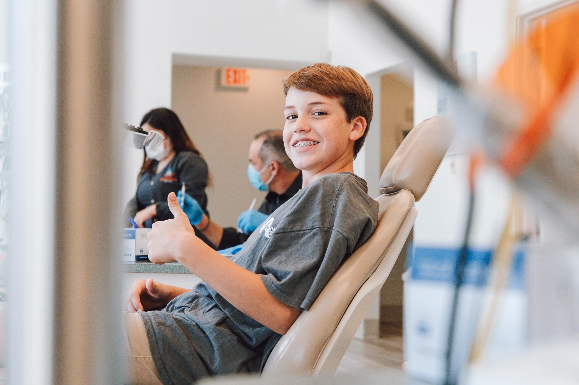 En pojke som sitter på en stol hos tandläkaren och gör tummen upp med ena handen.