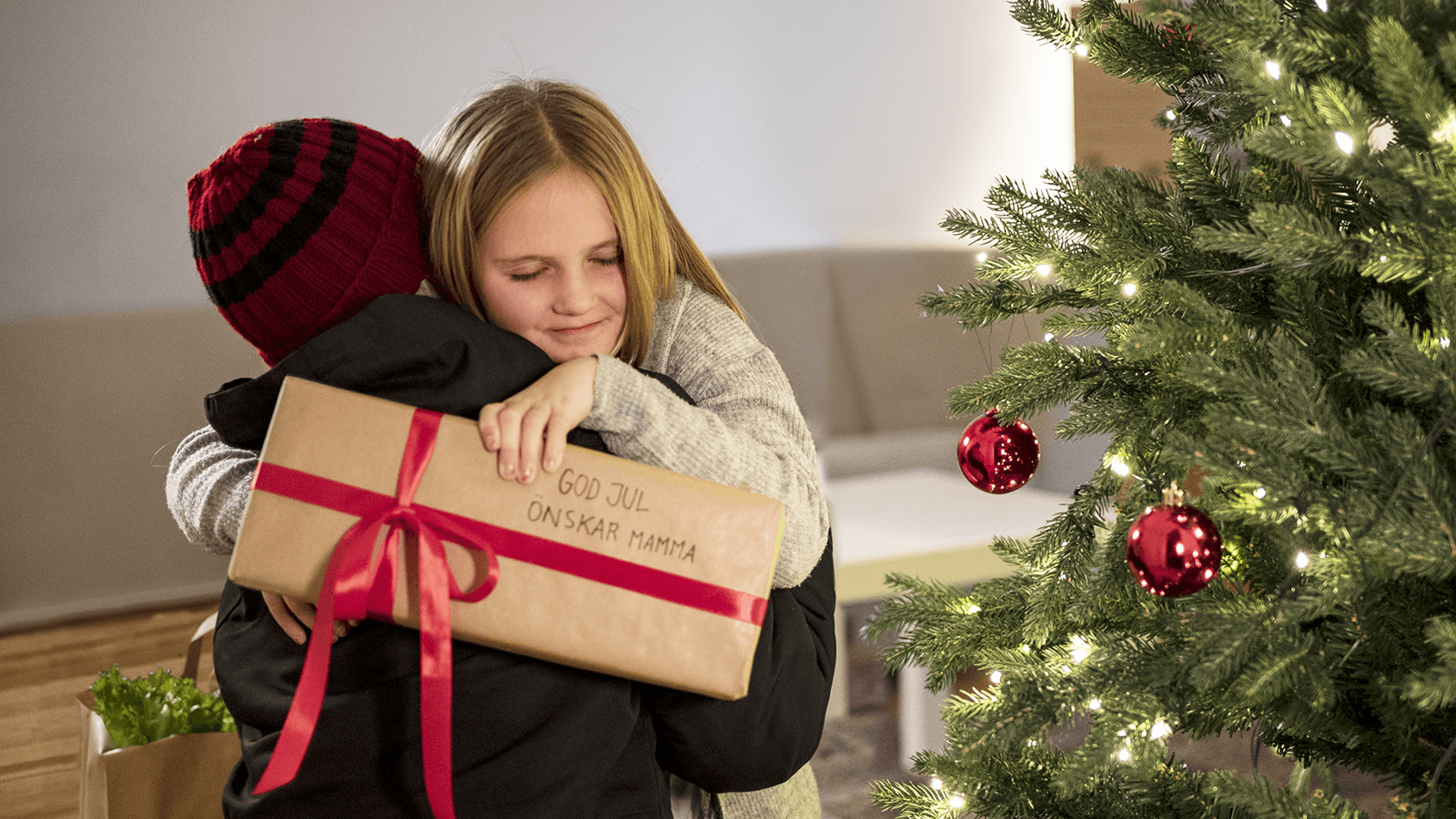 En mamma kramar om sin dotter bredvid en julgran, flickan håller i ett paket och en kasse mat står vid deras sida. 