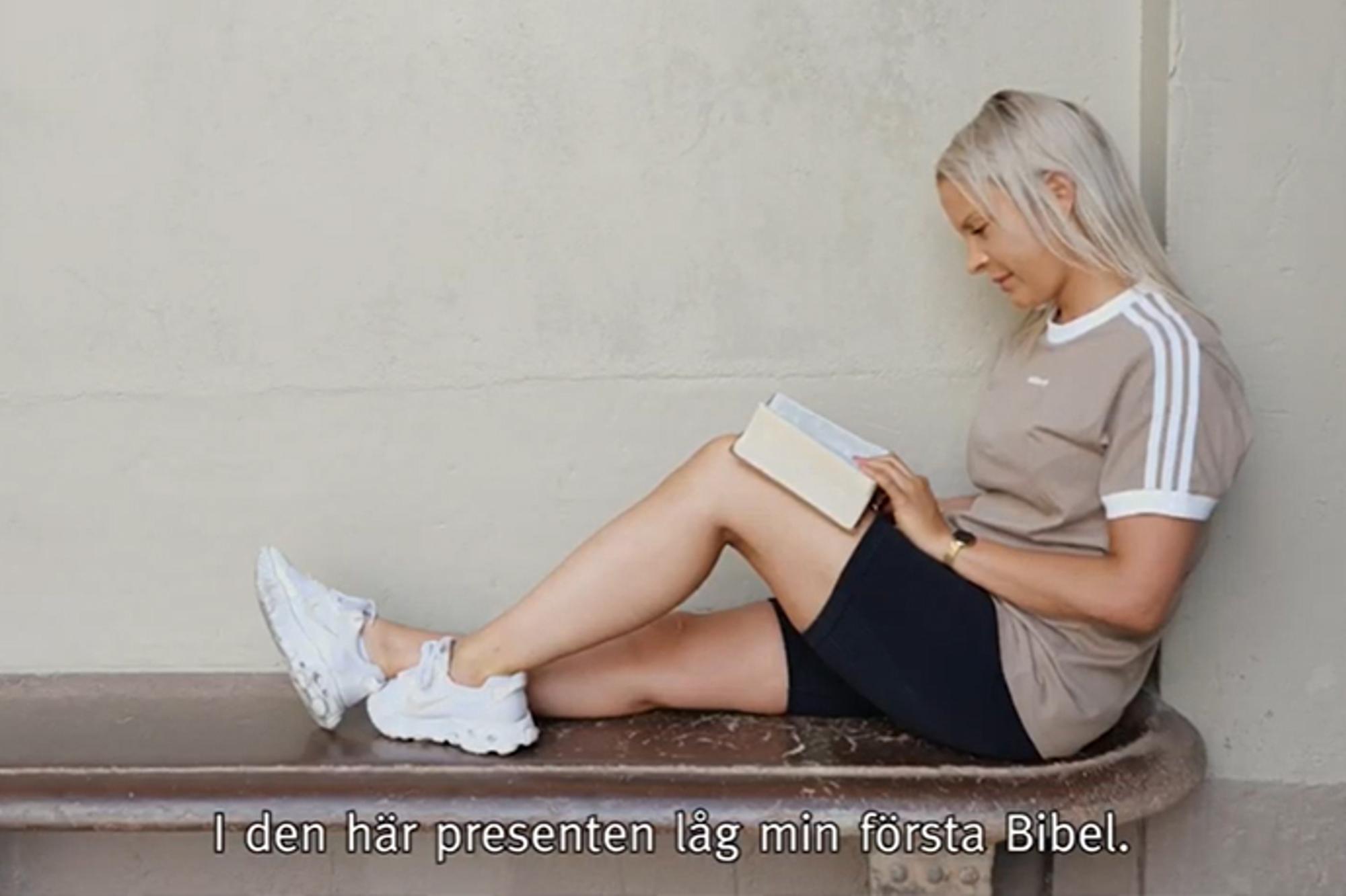 Carolina sitter på en bänk med ryggen lutad mot en vägg. I knät har hon en uppslagen bibel som hon läser i.