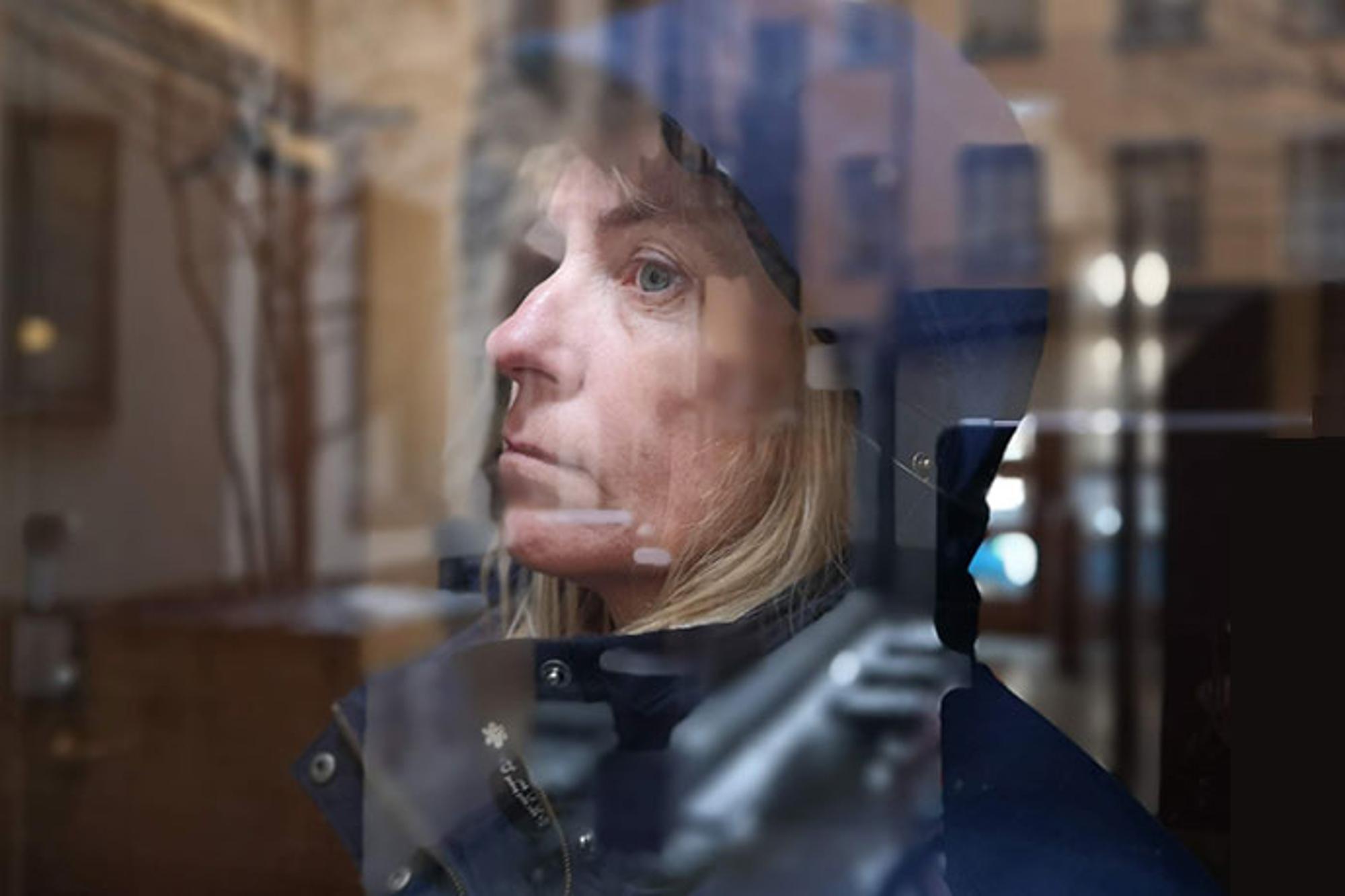 En kvinna som tittar ut genom ett fönster. Hon är iklädd en blå jacka med luva.